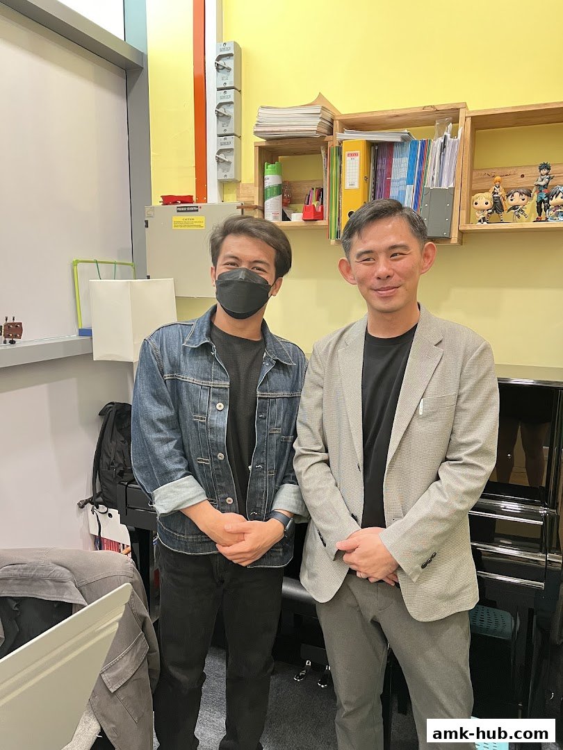 Two men stand in front of a piano, with shelves of books and figurines in the background.
