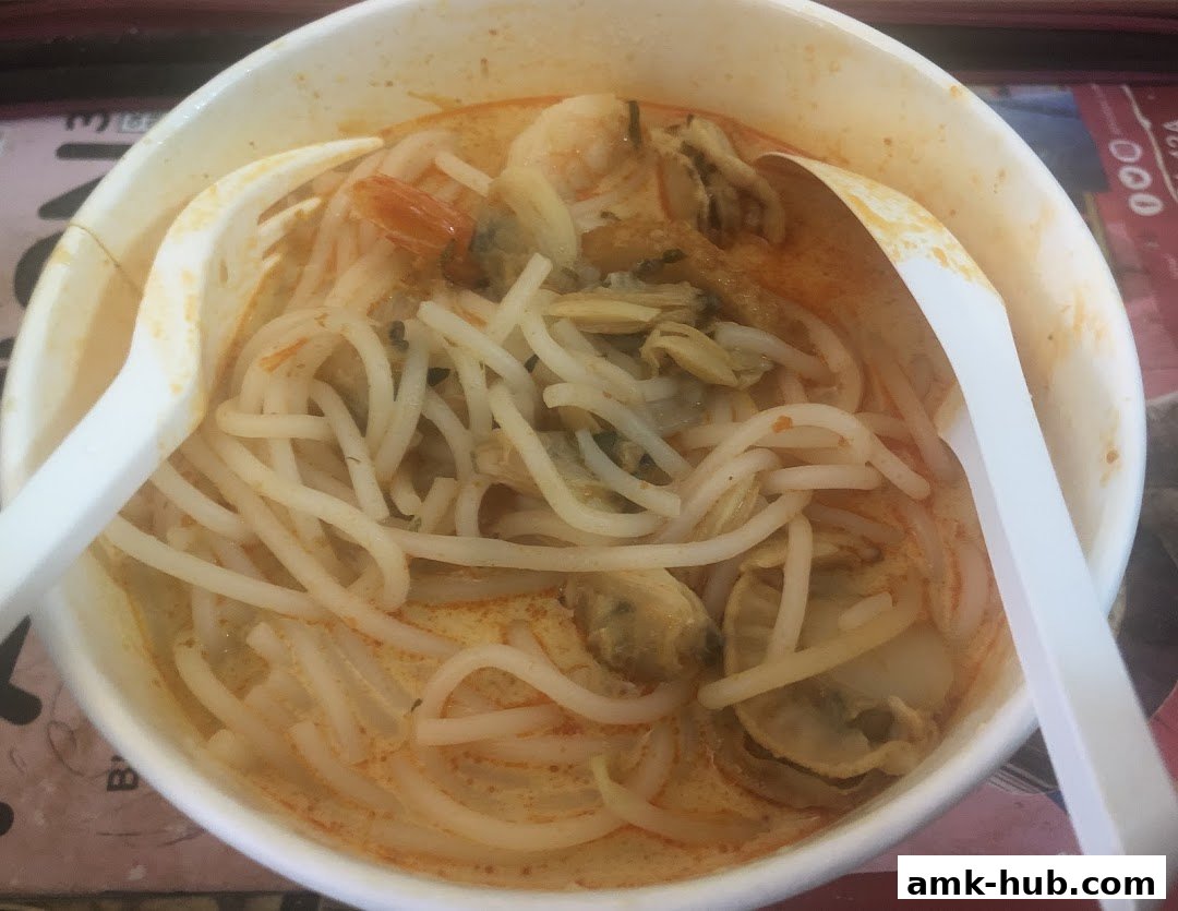 A close-up shot of a bowl of Laksa with noodles, shrimp, and clams, served with plastic cutlery.