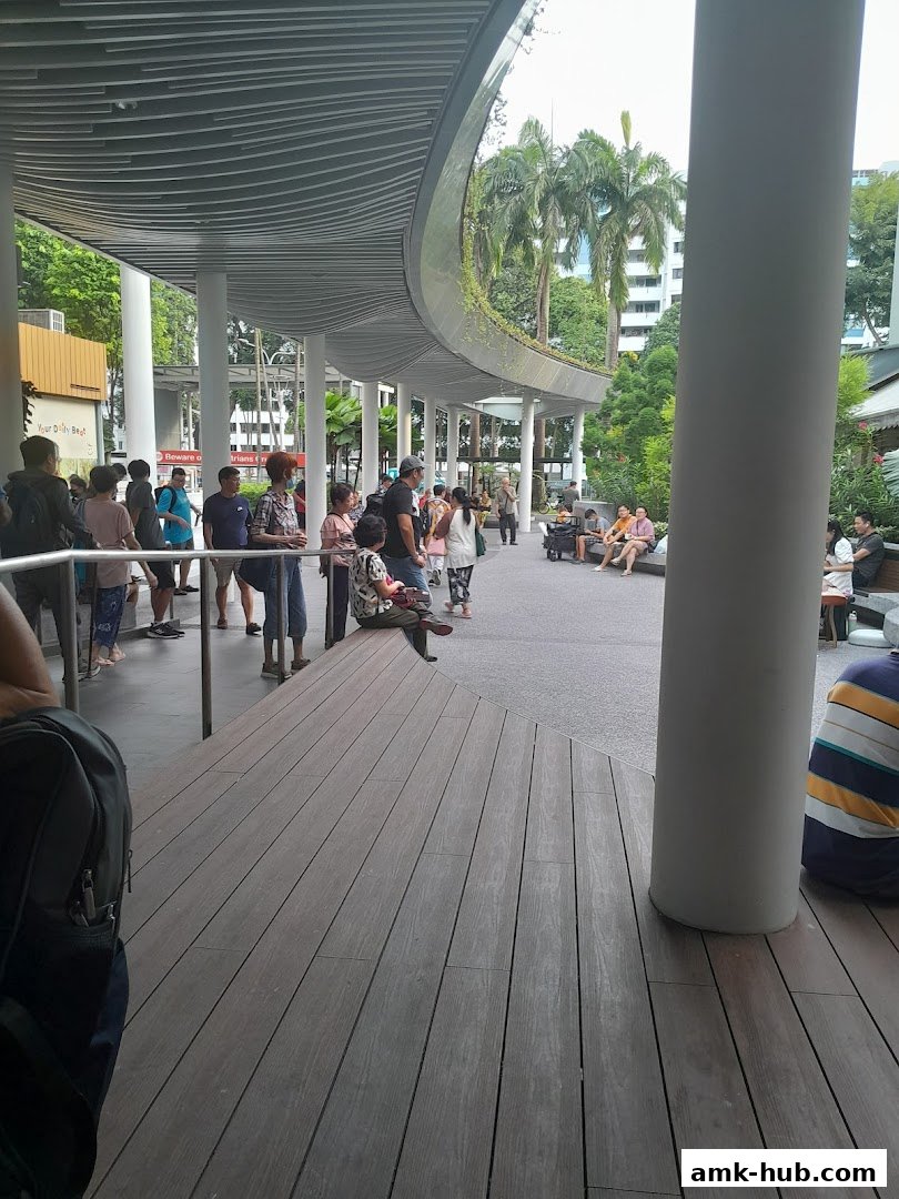 People gathered under a modern curved walkway with greenery and buildings in the background.