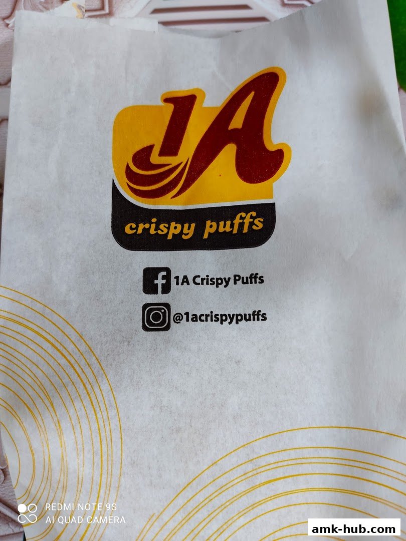 A white paper bag with the '1A Crispy Puffs' logo and social media handles.