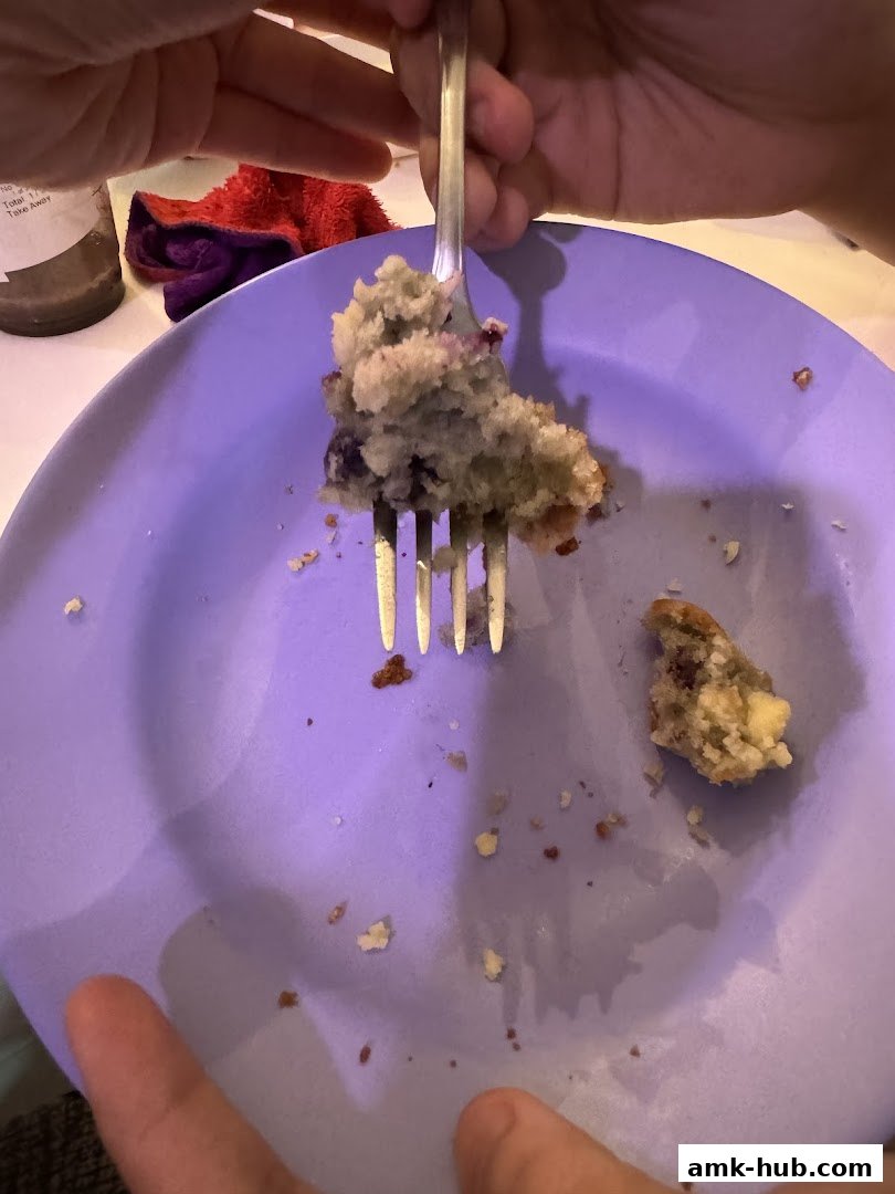 A fork lifts a piece of blueberry muffin from a purple plate with crumbs.