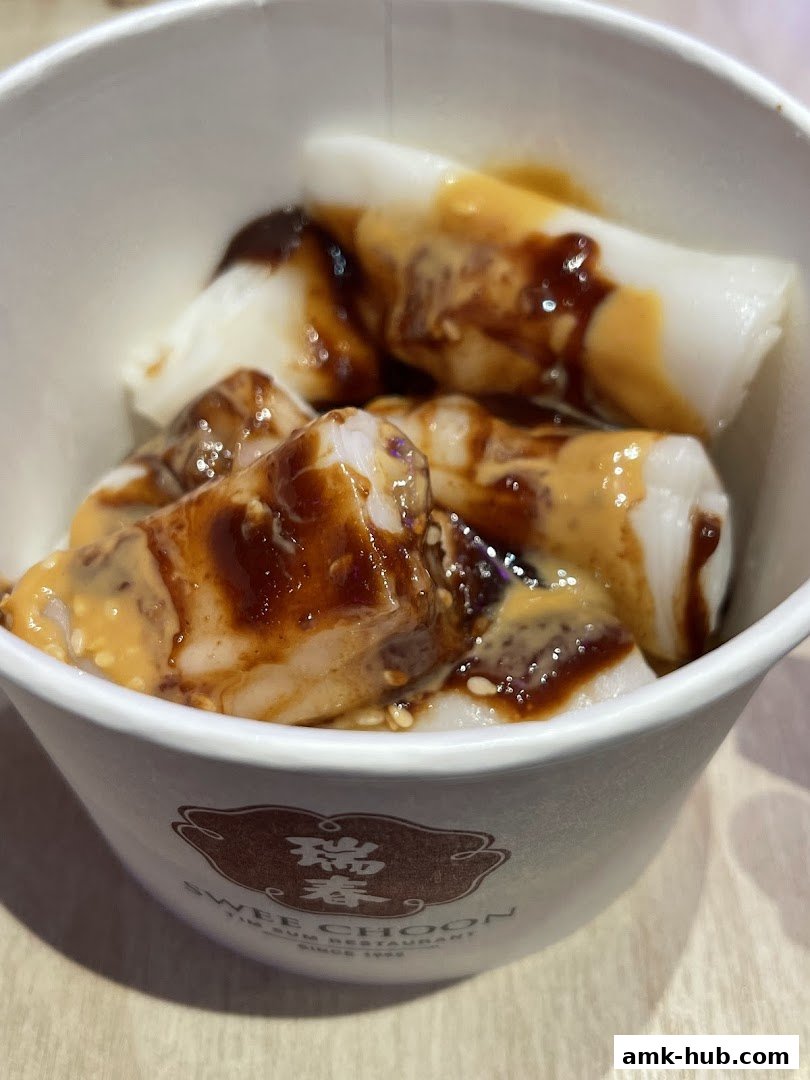 A close-up of dim sum rice noodle rolls drizzled with sauce and sesame seeds in a paper cup.