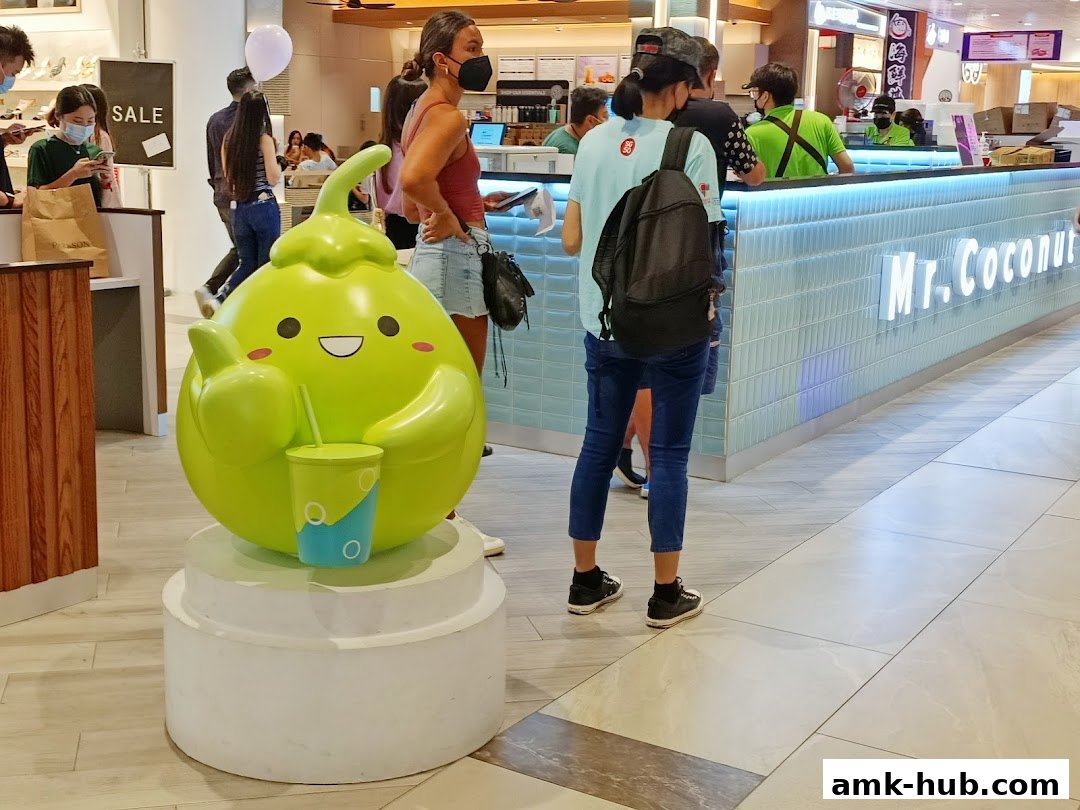 A cute coconut mascot stands in front of the Mr. Coconut shop counter with customers.