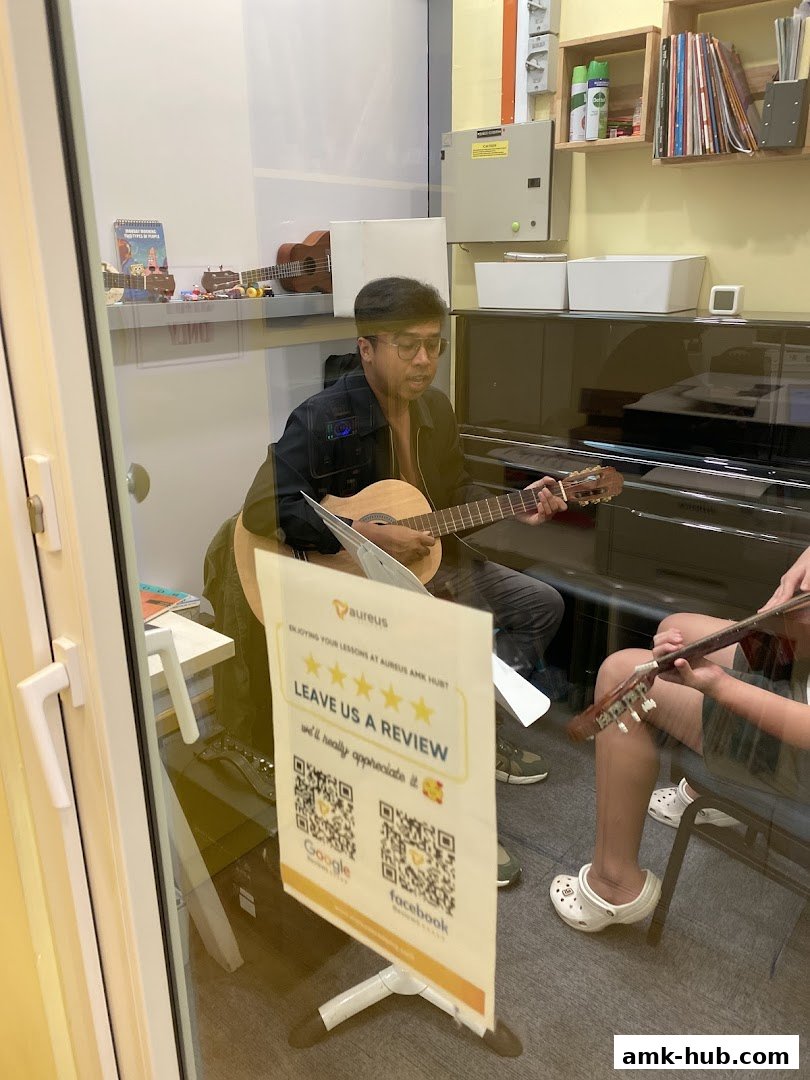 A man plays guitar in a music lesson room with a piano and ukuleles visible.