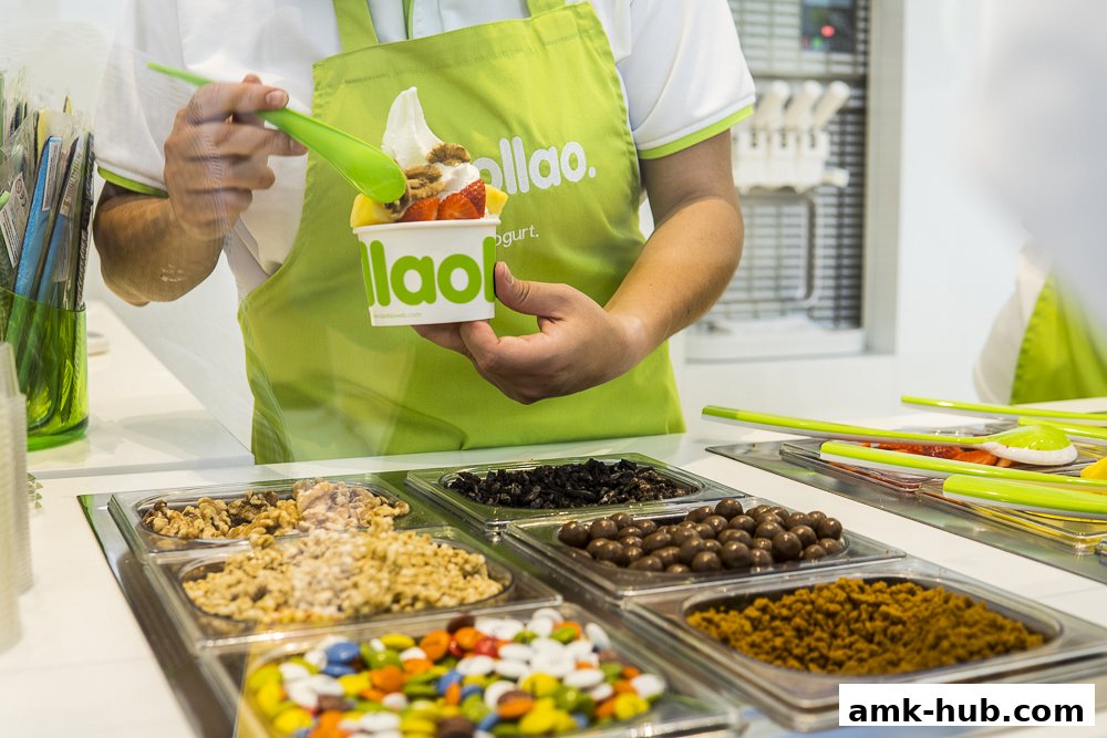 A person in a green apron prepares a frozen yogurt cup with fruit and toppings.
