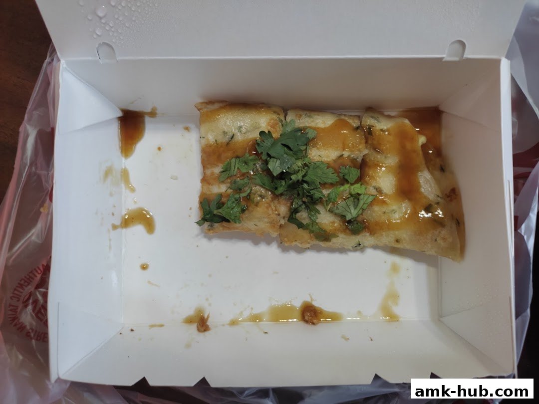 A piece of Taiwanese street food, likely oyster omelette, in a white takeout box with sauce and cilantro.