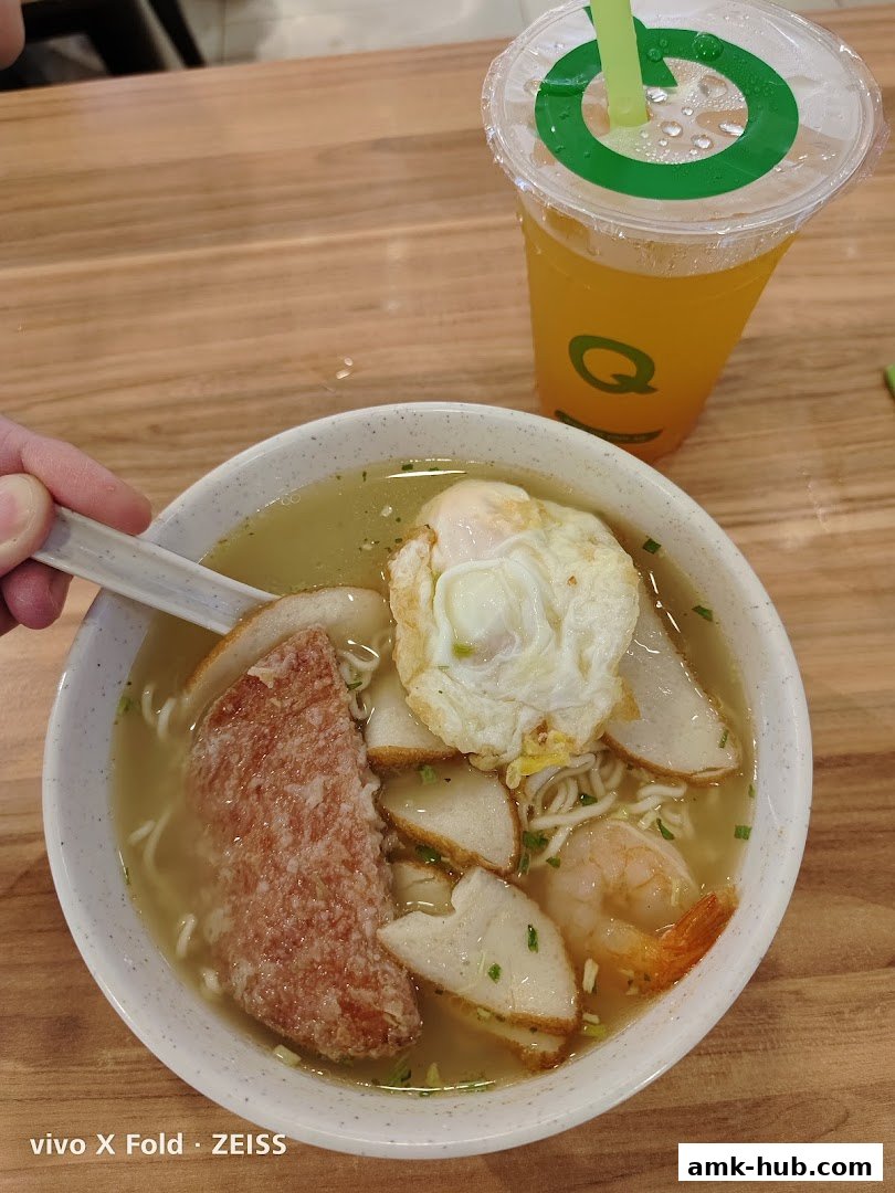 A bowl of noodle soup with fried meat, fish cakes, egg, and shrimp, next to a drink.