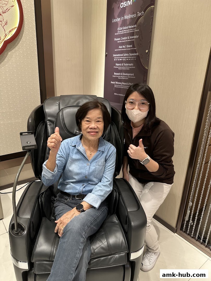 A woman sits in a massage chair, giving a thumbs up, with a staff member beside her.