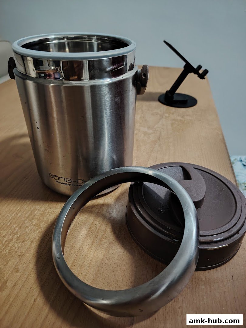 A stainless steel insulated container with its lid and a metal ring on a wooden surface.