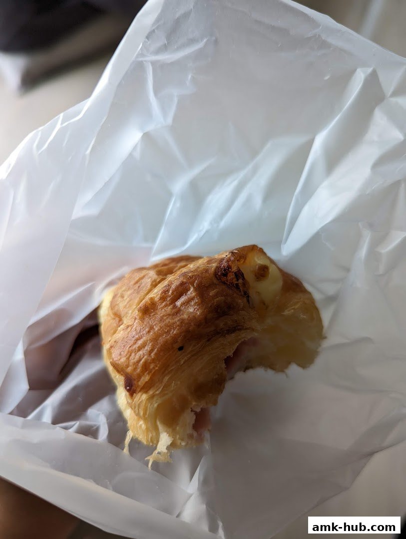 A close-up of a partially eaten ham and cheese pastry in a white paper bag.