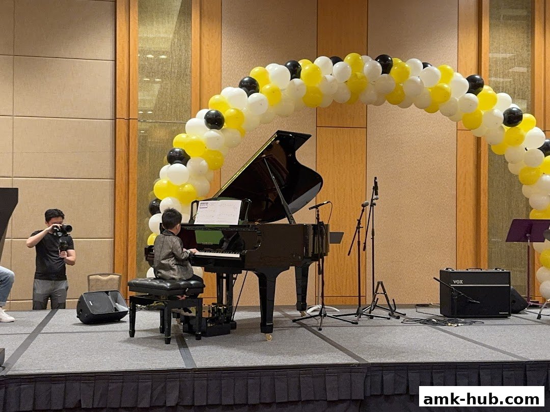 A young boy plays a grand piano on stage, filmed by a cameraman, under a balloon arch.