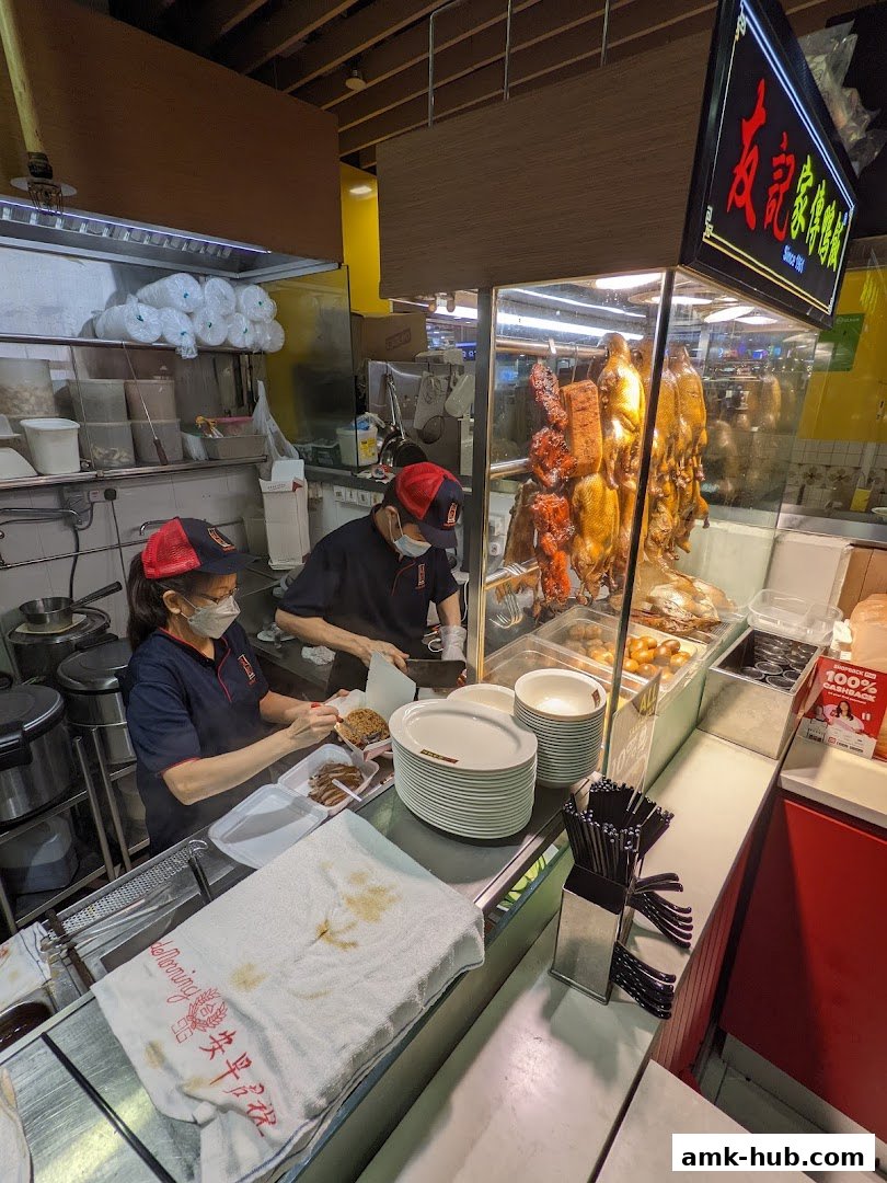 Staff preparing food behind a counter with roasted ducks and other meats on display.