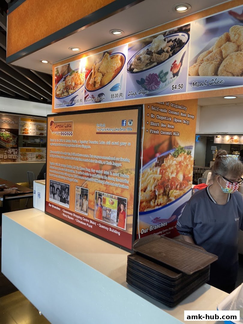 A food stall counter with a menu board displaying Laksa and other dishes, and a staff member.