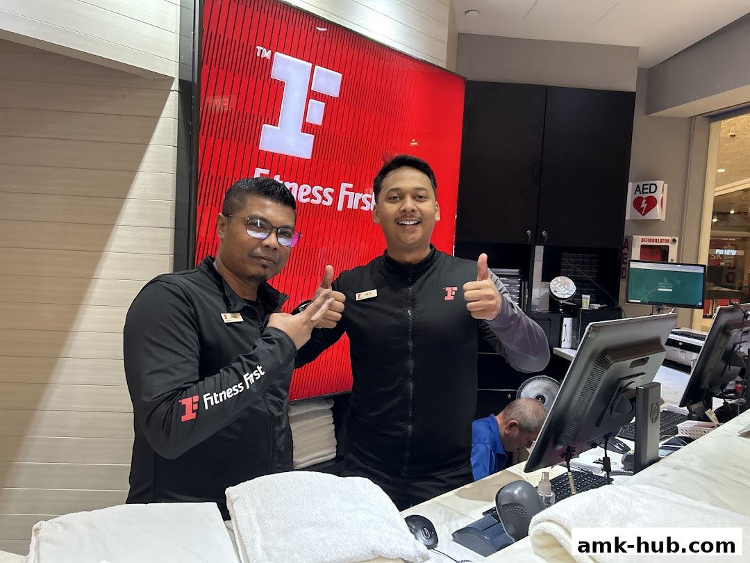 Two smiling staff members give thumbs up in front of the Fitness First logo and reception desk.