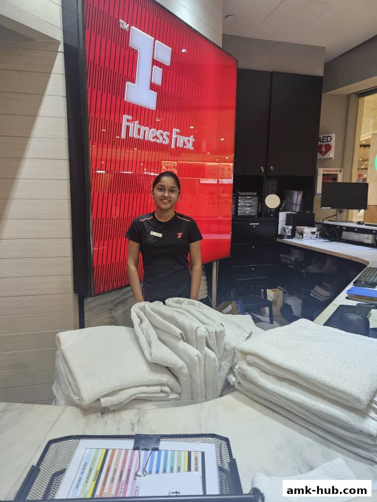 A smiling staff member stands behind a counter with folded towels at Fitness First.