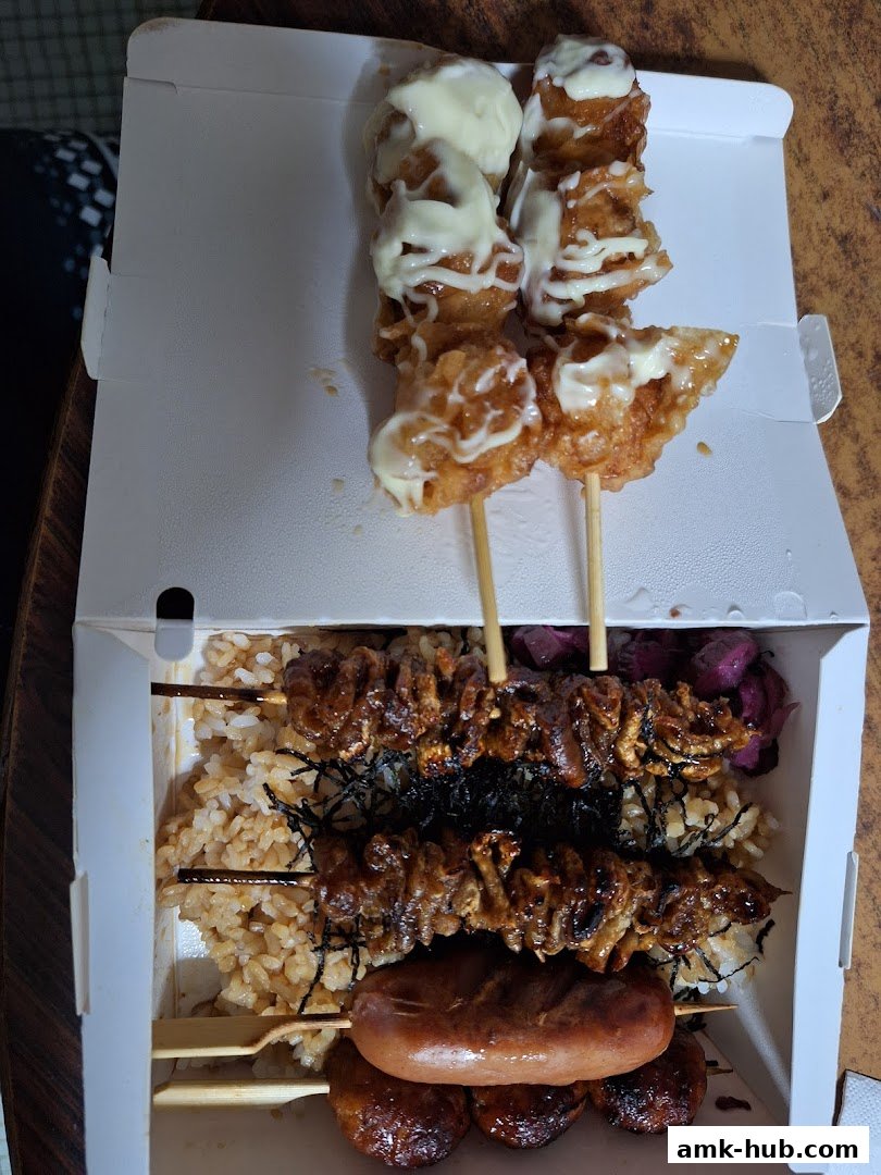 A takeout box filled with assorted skewers of grilled meats, rice, and a sausage.