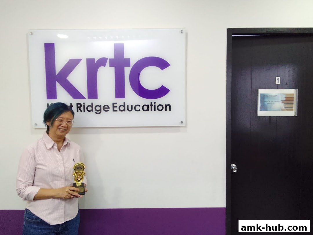 A woman holds a trophy in front of the KRTC Ang Mo Kio Education sign.