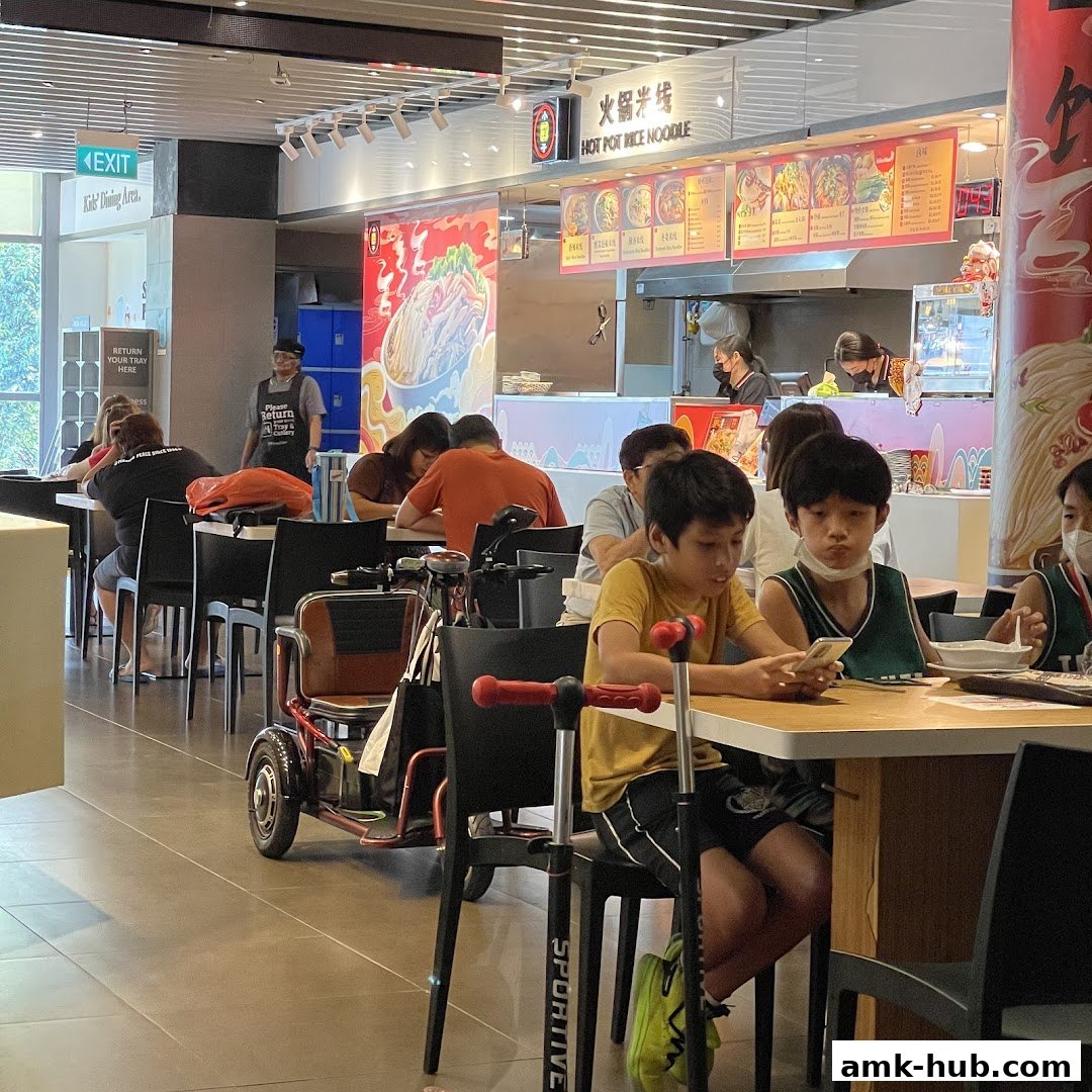People dining at tables in a food court with a hot pot noodle stall in the background.