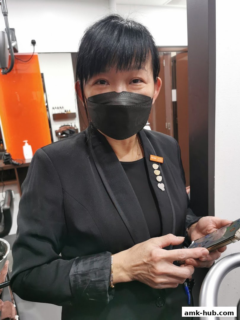 A woman wearing a black blazer and mask stands in a salon, holding a phone.