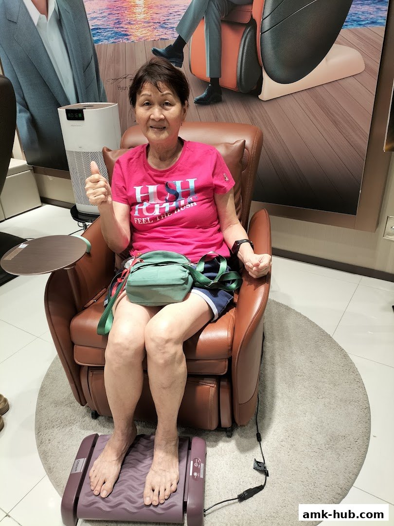 An elderly woman sits in a massage chair, giving a thumbs-up, with a foot massager.