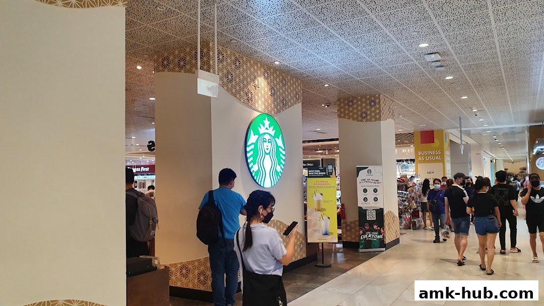 People walk past the iconic Starbucks logo and a promotional poster inside a mall.