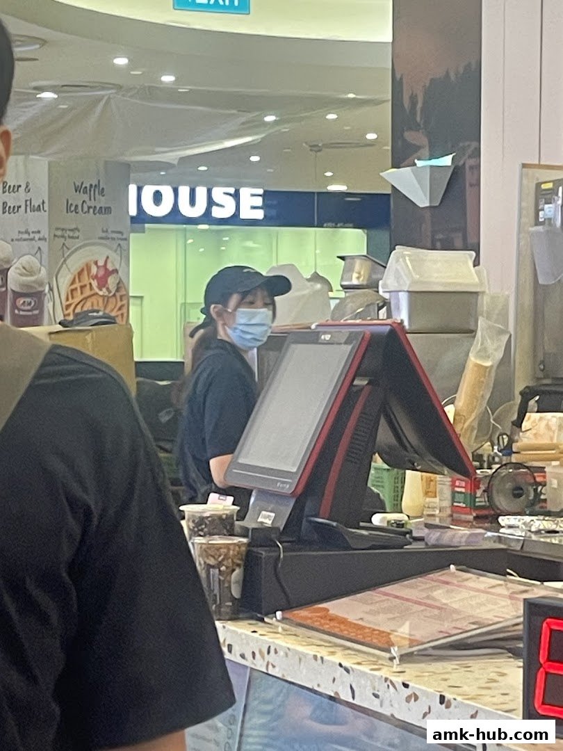 A staff member wearing a mask serves customers at a counter with a POS system.