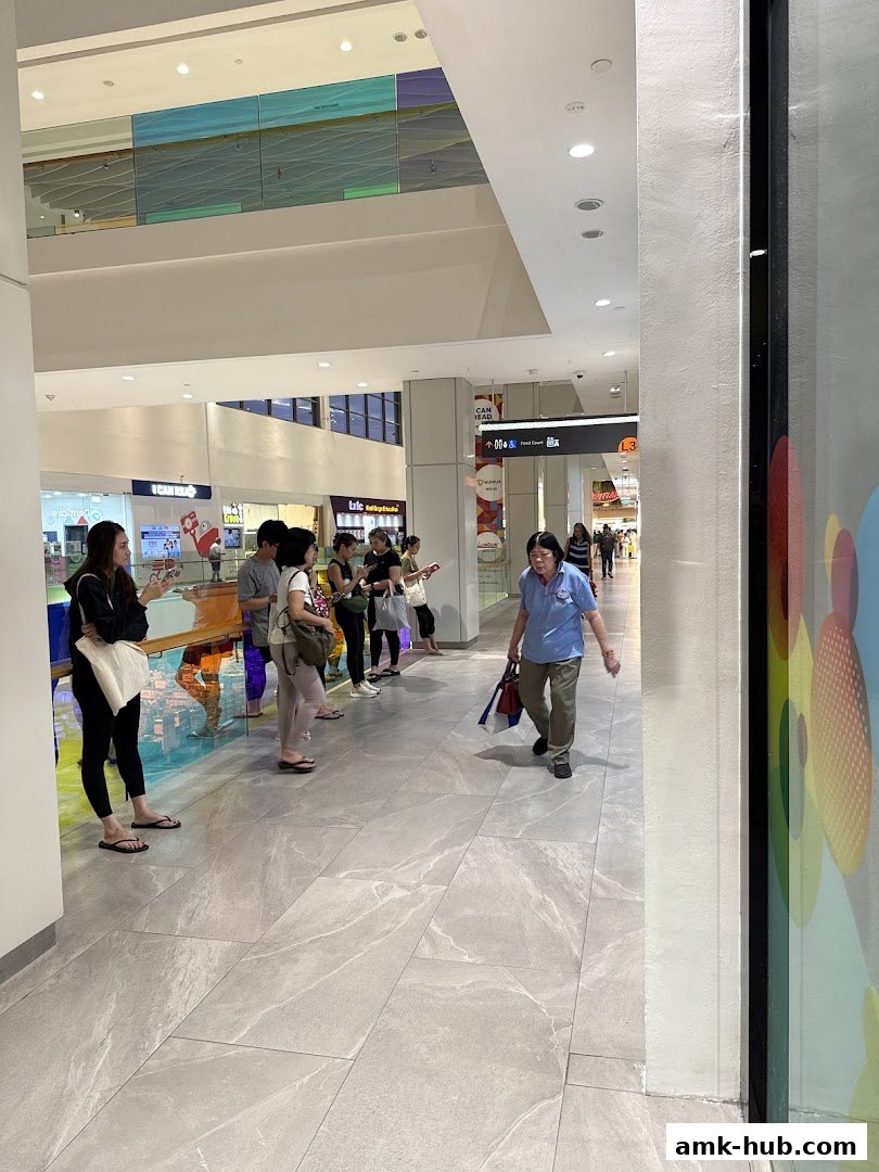People walking and standing in a modern shopping mall hallway with colorful art on the ceiling.