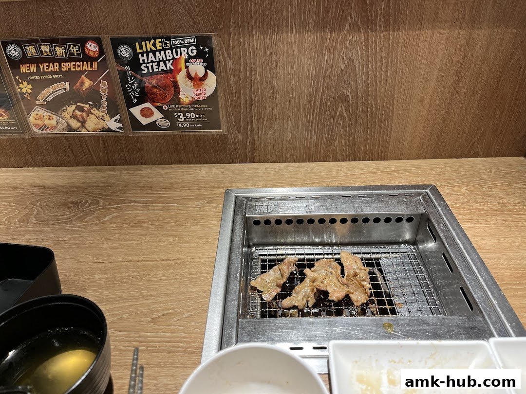Meat grilling on a barbecue at Yakiniku Like AMK Hub with New Year specials advertised.