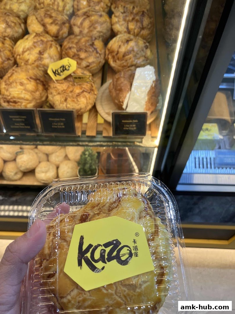 A hand holds a Kazo pastry in a clear container, with a display of pastries in the background.