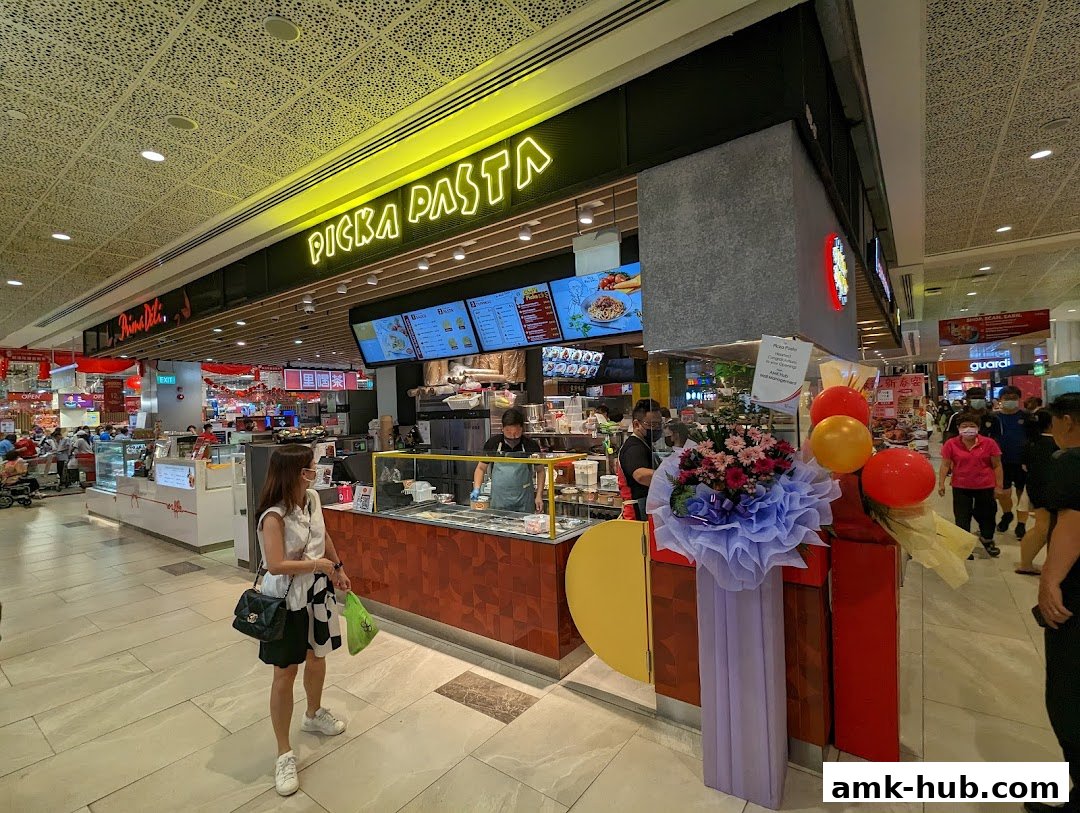A woman stands outside the brightly lit 'Picka Pasta' shop with a menu displayed.