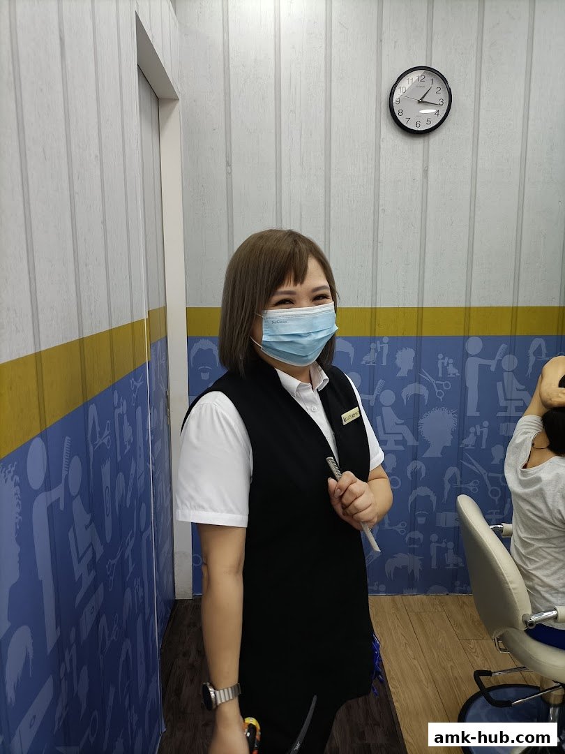 A smiling hairstylist wearing a mask and vest stands in a salon with a clock on the wall.