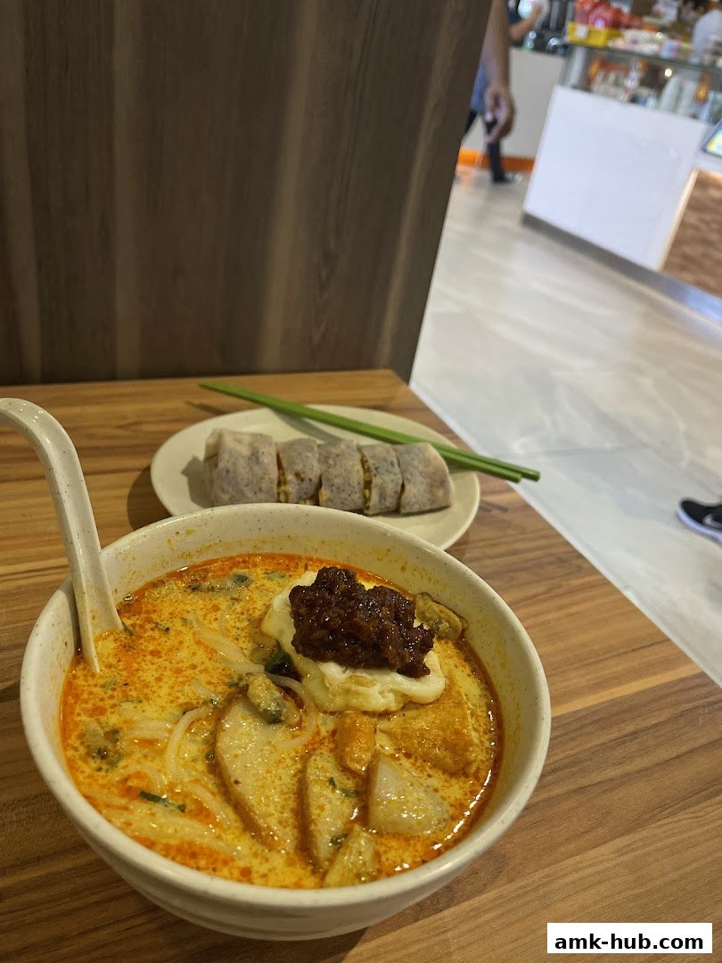 A bowl of Laksa with a side of popiah on a wooden table.