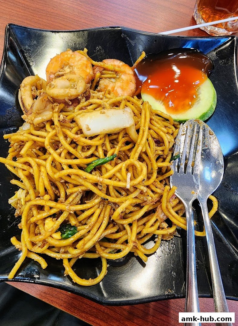 A plate of stir-fried noodles with seafood, sauce, and a slice of cucumber.