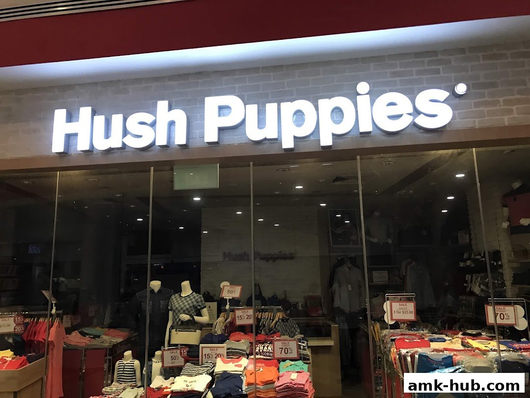 The exterior of a Hush Puppies store with a prominent illuminated sign and displays of clothing.