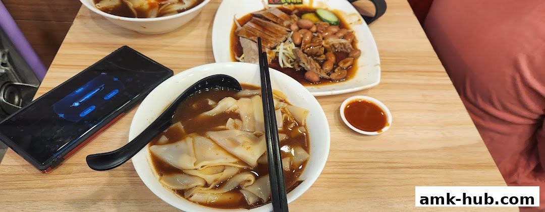 A close-up of a table with bowls of noodles, roasted duck, and a small dish of sauce.