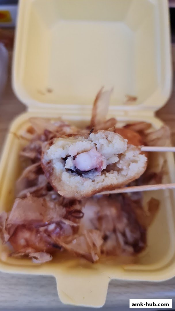 A close-up of a takoyaki ball being held by chopsticks, revealing the octopus inside.