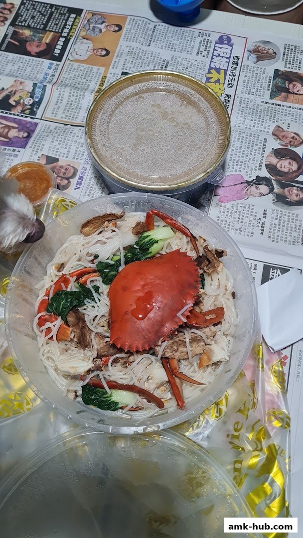 A close-up of a crab noodle dish with vegetables and a side of soup, presented on newspapers.