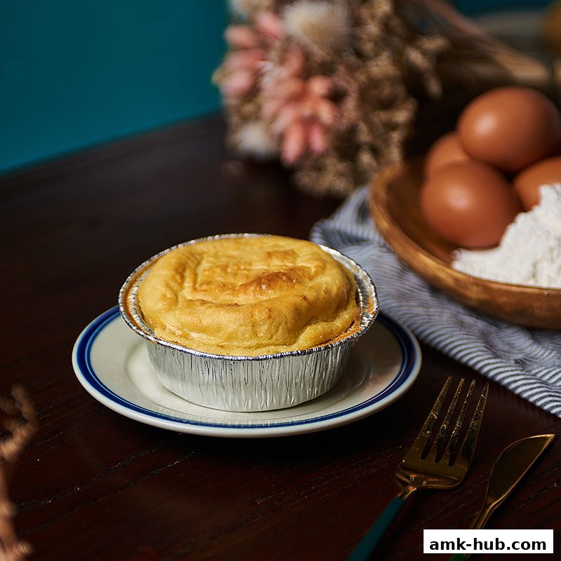A golden-brown chicken pie in a foil container on a plate, with eggs and flour in the background.