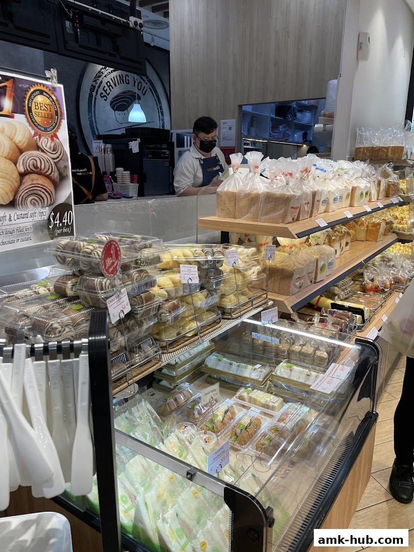 A bakery display case filled with various breads, pastries, and sandwiches, with a staff member behind the counter.