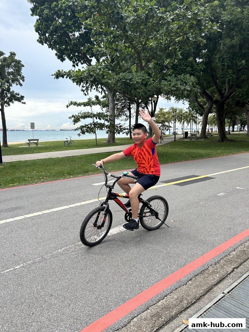 A young boy waves while riding a bicycle on a paved path by the sea.