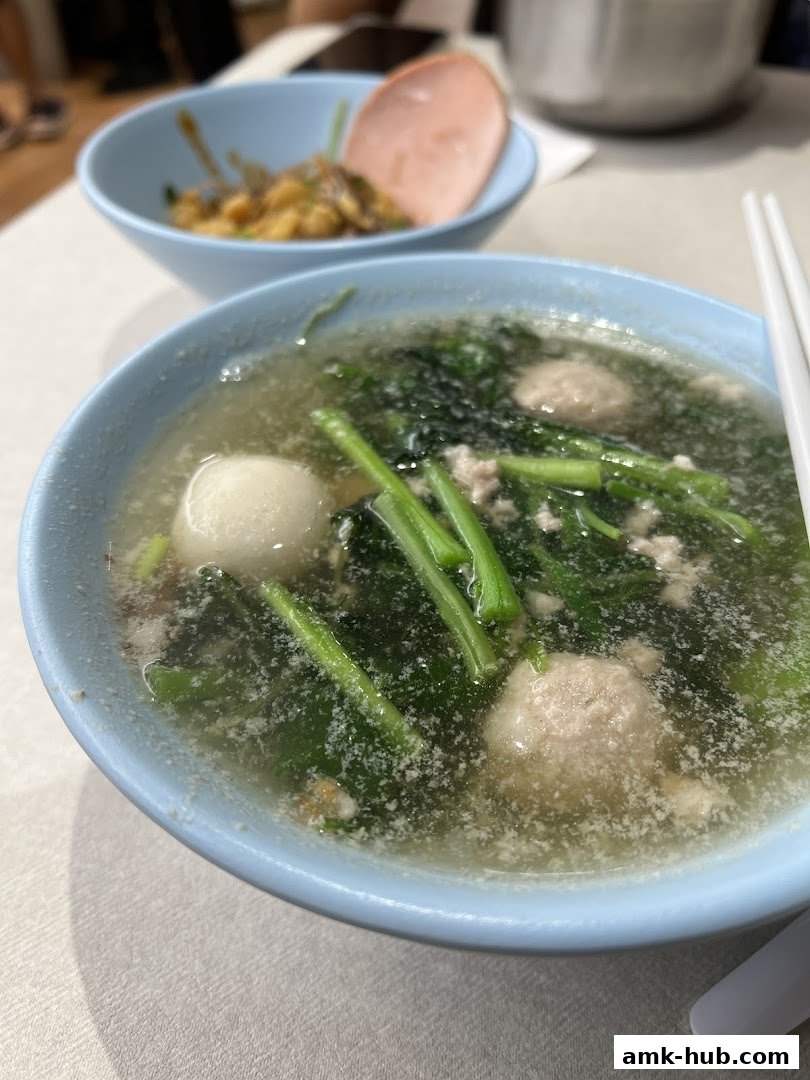 A close-up of a bowl of soup with meatballs and greens, with another dish in the background.
