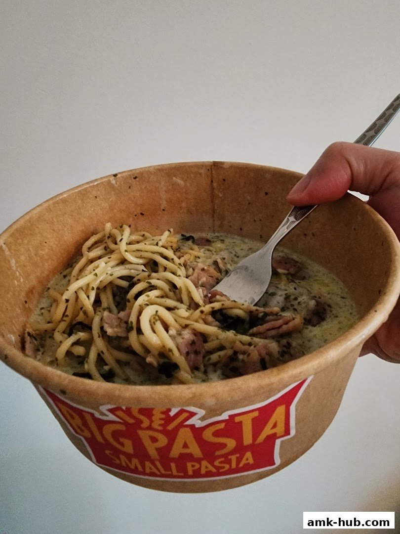 A hand holds a bowl of spaghetti with bacon and creamy sauce, with the shop's logo visible.