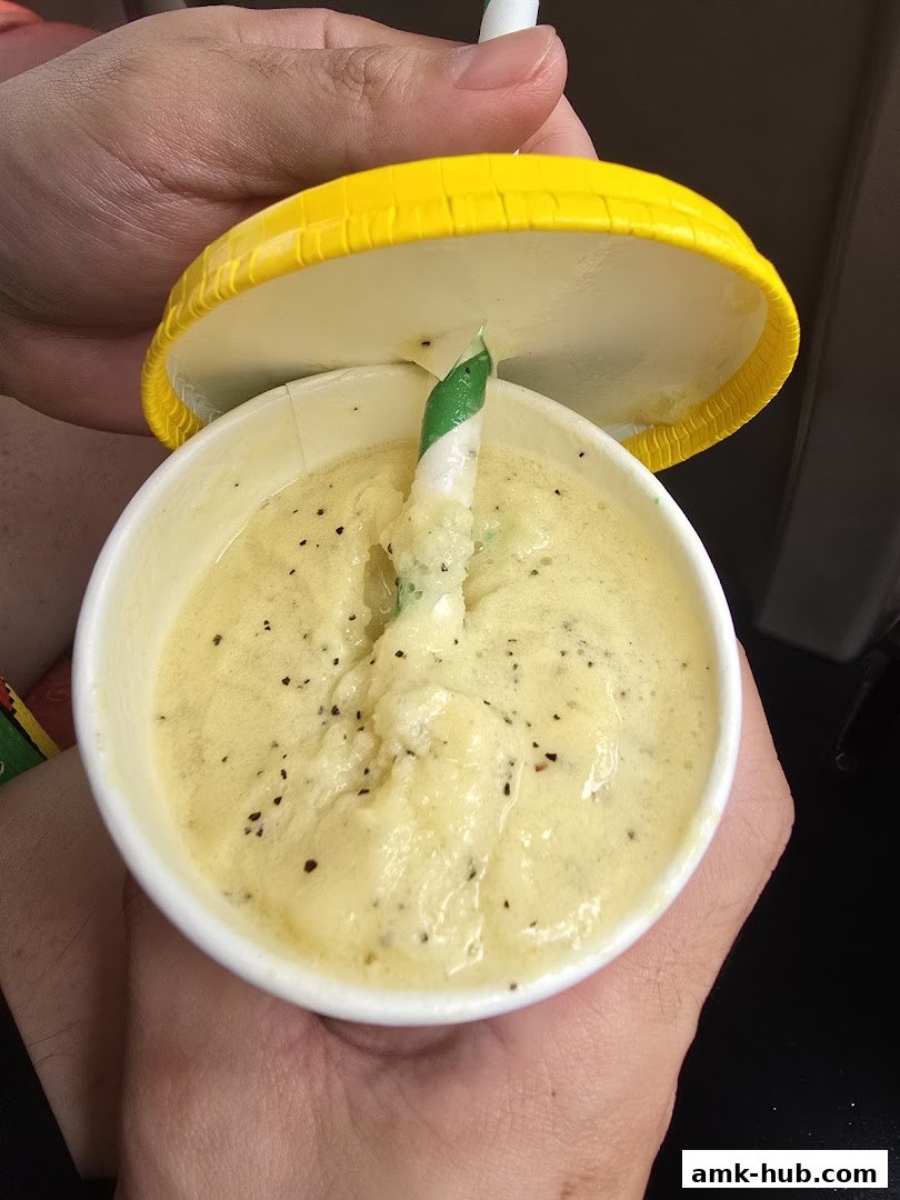 Hands holding a cup with a yellow lid, containing a yellow smoothie with a striped straw.