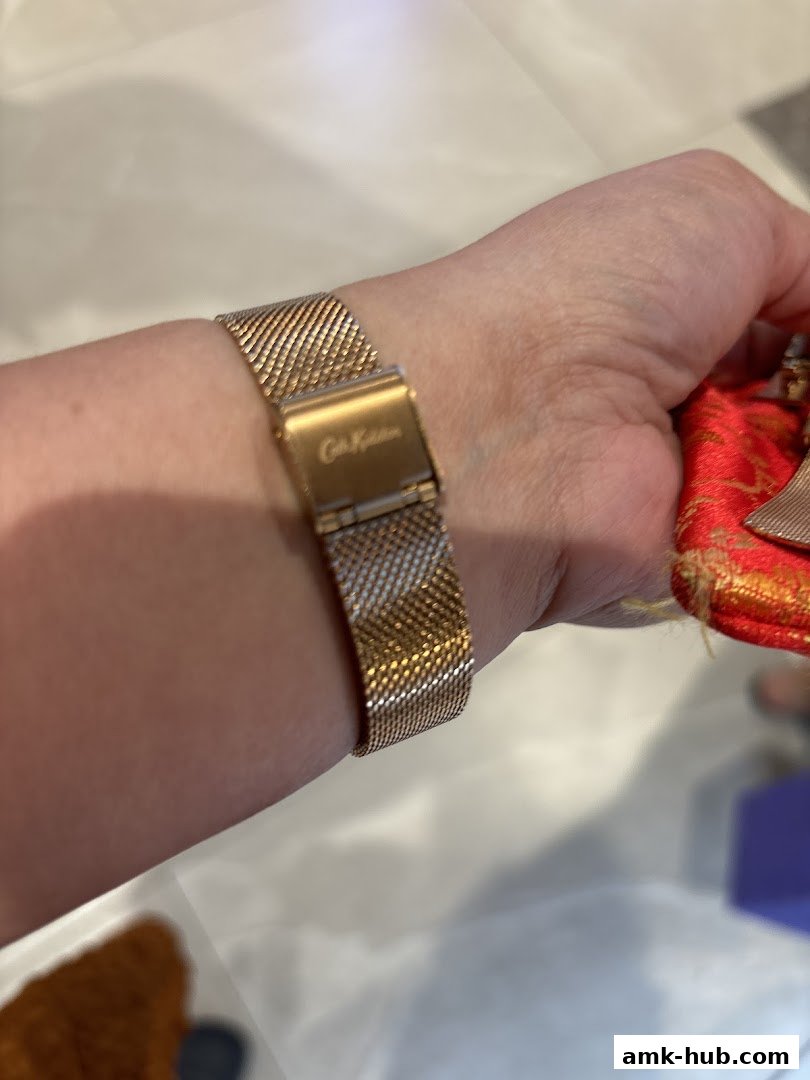A person's wrist wearing a rose gold mesh watch with a branded clasp.