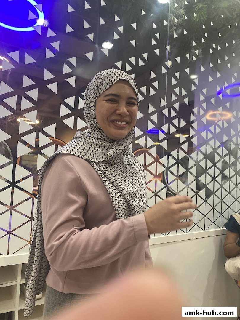 A smiling woman wearing a hijab and a pink blazer stands in front of a patterned glass wall.