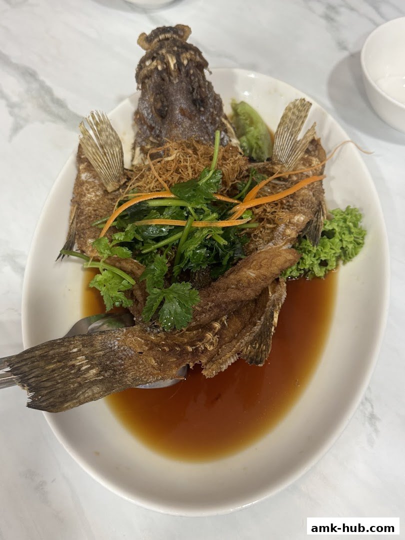 A whole fried fish served with sauce, herbs, and shredded vegetables on a white plate.