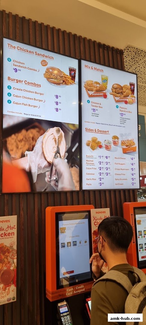 A digital menu board displays Popeyes chicken sandwich combos, burger combos, and sides with prices.