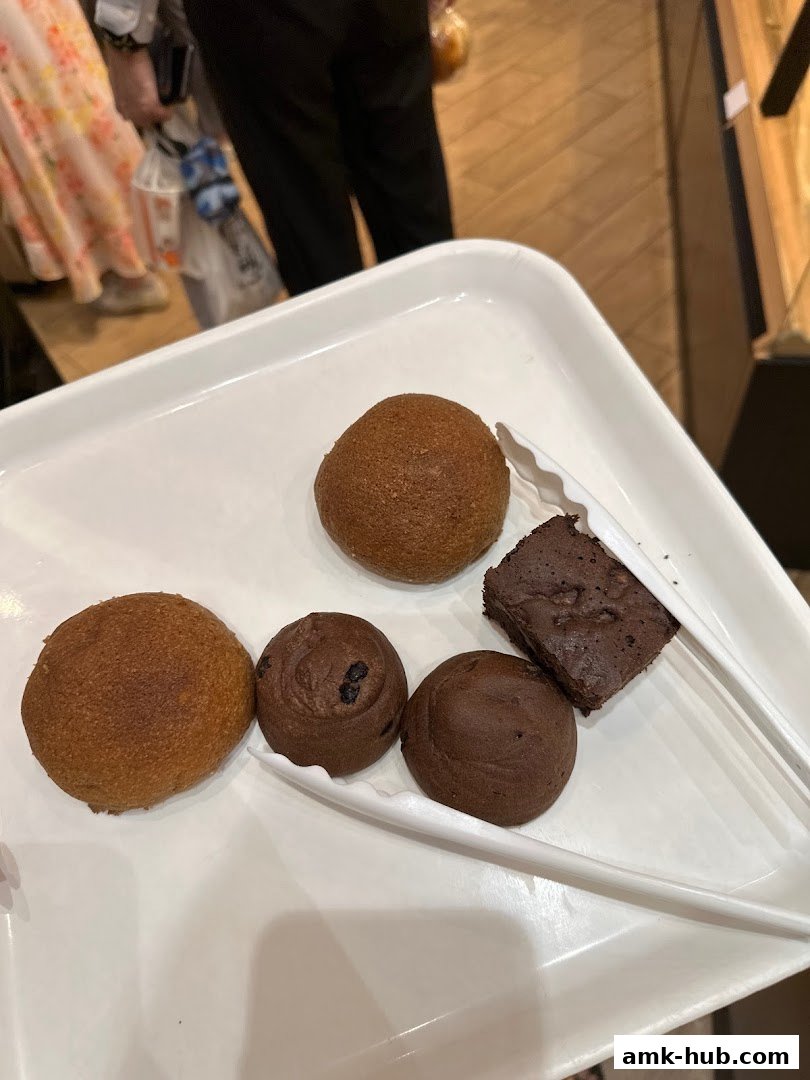 A white tray holds assorted baked goods, including buns and a brownie, with tongs.