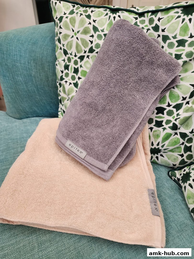 Two soft epitex towels, one beige and one grey, are displayed on a teal couch with patterned cushions.