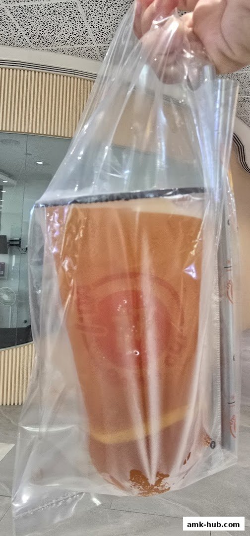A hand holds a clear plastic bag containing a large beverage with an orange hue.